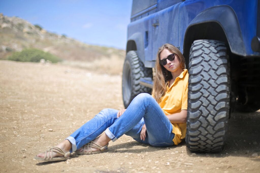 A woman wearing sunglasses relaxes against a blue off-road vehicle in a desert landscape.